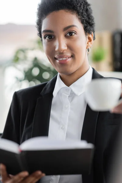 Felice manager afroamericano in blazer nero guardando la fotocamera vicino alla tazza di caffè offuscata e notebook in ufficio — Foto stock