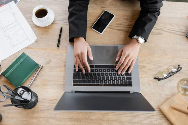 Вид сверху на срезанный африканский американский менеджер, печатающий на ноутбуке рядом со смартфоном и кофейной чашкой на рабочем столе в офисе — стоковое фото