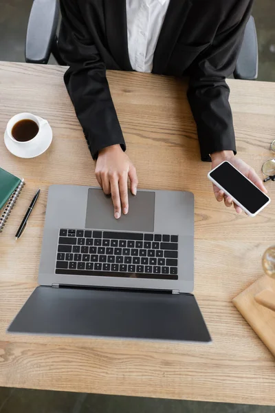 Вид зверху обрізаної афроамериканської бізнес-леді, використовуючи ноутбук і тримаючи смартфон з порожнім екраном біля чашки кави на столі в офісі — Stock Photo
