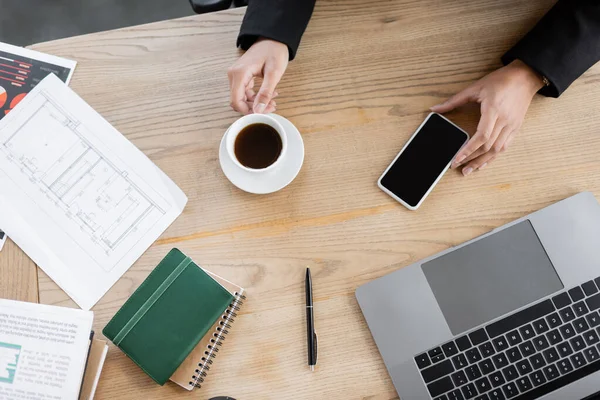 Recortado de Africano americano empresária segurando smartphone com tela em branco e xícara de café perto de laptop na mesa de trabalho no escritório — Fotografia de Stock