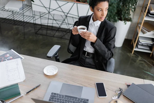Vista de ángulo alto de la pensativa mujer de negocios afroamericana en chaqueta negra sentada con taza de café y mirando hacia otro lado cerca del escritorio de trabajo en la oficina - foto de stock