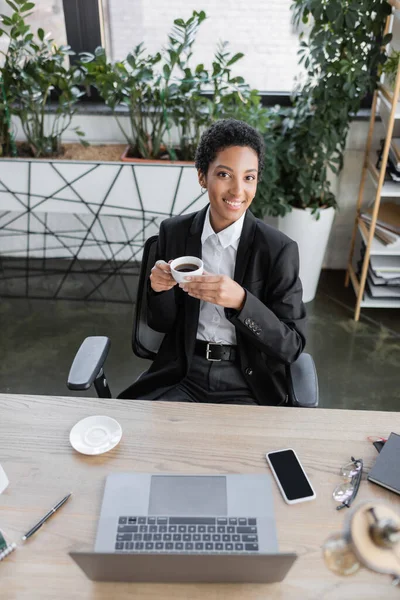 Vista ad alto angolo di successo afro-americana donna d'affari sorridente alla macchina fotografica mentre seduto con tazza di caffè vicino gadget — Foto stock