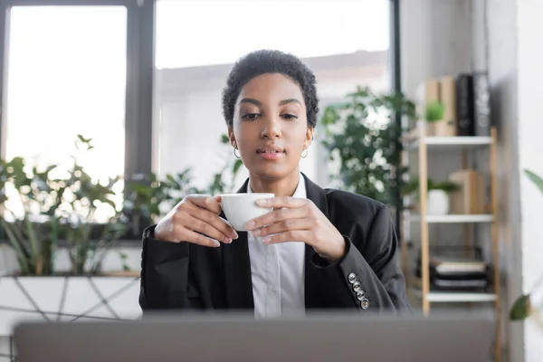 Стильна афроамериканська бізнесменка в чорному блейзері, що тримає чашку кави і дивиться на розмитий ноутбук в офісі — стокове фото