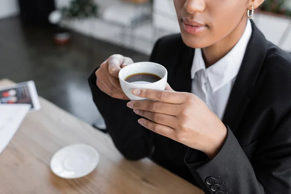 Частковий вид на афроамериканську ділову жінку в чорному блейзері сидячи з чашкою чорної кави на офісі — стокове фото