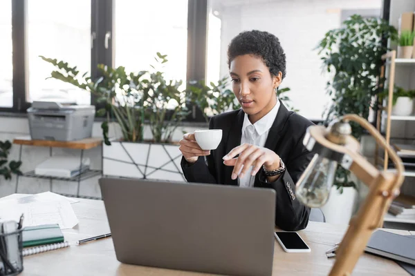 Jovem empresária afro-americana com xícara de café apontando para laptop perto de smartphone e lâmpada de mesa borrada no escritório moderno — Fotografia de Stock