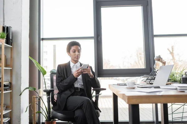 Élégante femme d'affaires afro-américaine messagerie sur téléphone mobile près d'un ordinateur portable sur le bureau dans un bureau moderne — Photo de stock