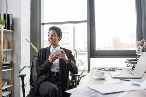 Donna d'affari afro-americana di successo con smartphone che distoglie lo sguardo mentre si siede vicino alla scrivania offuscata in ufficio — Foto stock