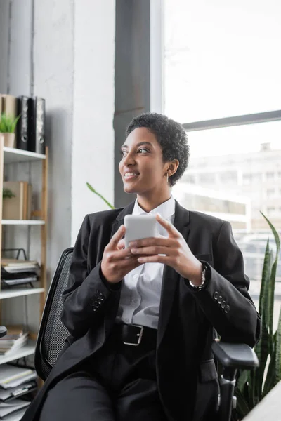 Felice donna d'affari africana americana in tuta nera che tiene lo smartphone e distoglie lo sguardo mentre è seduta in ufficio — Foto stock