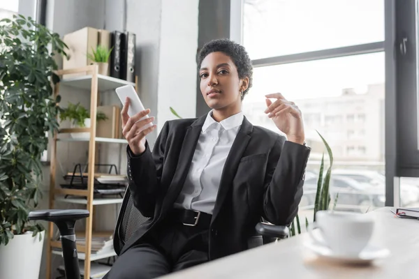 Junge afrikanisch-amerikanische Geschäftsfrau im schwarzen Anzug hält Handy in der Hand und blickt in die Kamera in der Nähe einer verschwommenen Kaffeetasse im Büro — Stockfoto