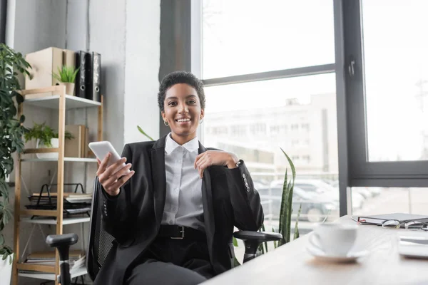 Alegre afroamericana mujer de negocios en elegante ropa formal celebración de teléfono celular y sonriendo a la cámara cerca borrosa taza de café en la oficina - foto de stock