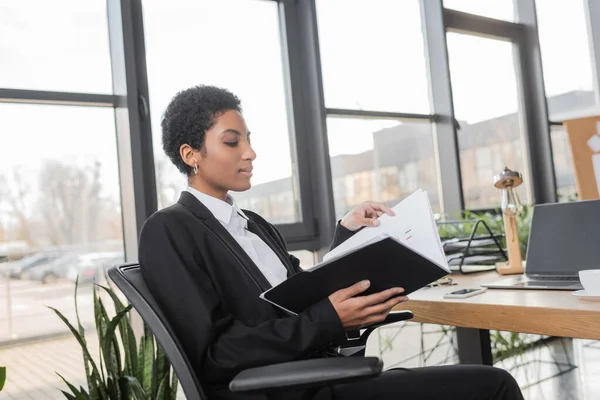 Vue latérale d'une femme d'affaires afro-américaine dans un blazer noir assis avec un dossier près d'un ordinateur portable avec un écran vide au bureau — Photo de stock