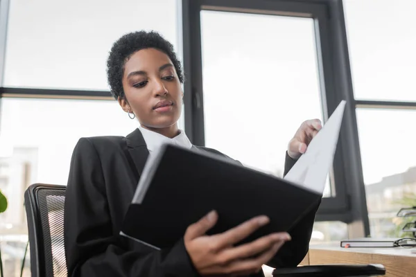 Vista de ángulo bajo de la joven mujer de negocios afroamericana que sostiene la carpeta con documentos mientras está sentada en la oficina - foto de stock
