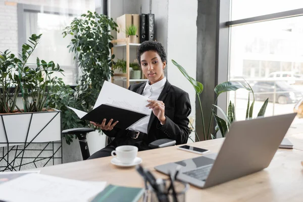Afrikanisch-amerikanische Geschäftsfrau hält Ordner mit Papieren in der Hand und blickt auf Laptop auf Schreibtisch in modernem Büro — Stockfoto
