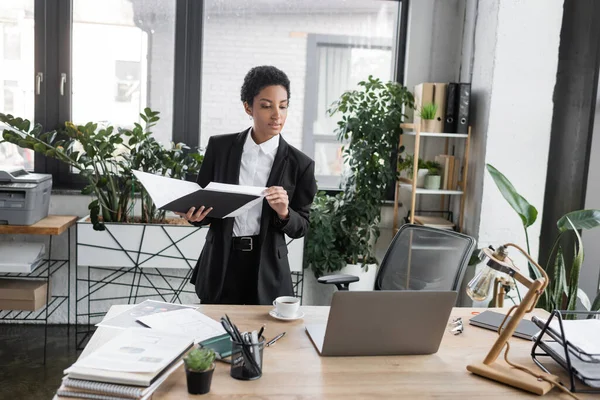 Афроамериканська бізнесменка в чорному костюмі тримає папку з документами і дивиться на ноутбук на робочому столі в сучасному офісі. — стокове фото