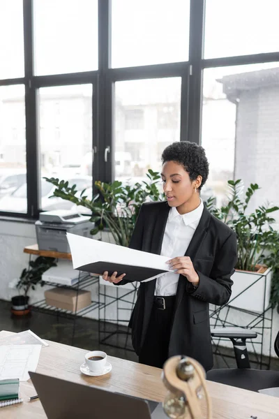 Élégante femme d'affaires afro-américaine tenant dossier avec des documents tout en se tenant près de tasse de café sur le bureau dans le bureau — Photo de stock