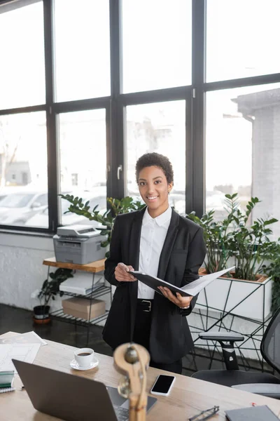Happy african american businesswoman holding folder with documents and looking at camera while standing near laptop in modern office — Stock Photo