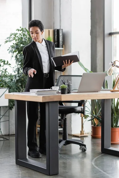 Стильна афроамериканська бізнес-леді, що стоїть з документами і вказує на блокноти біля ноутбука на робочому столі в офісі — стокове фото