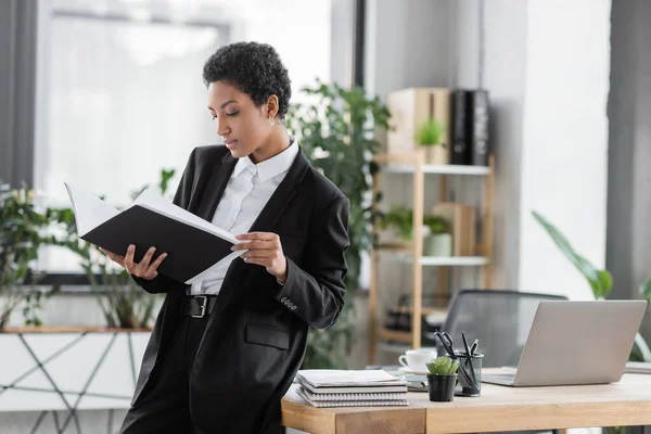 Joven mujer de negocios afroamericana mirando los documentos en la carpeta mientras está de pie cerca del escritorio con el ordenador portátil y los cuadernos en la oficina — Stock Photo