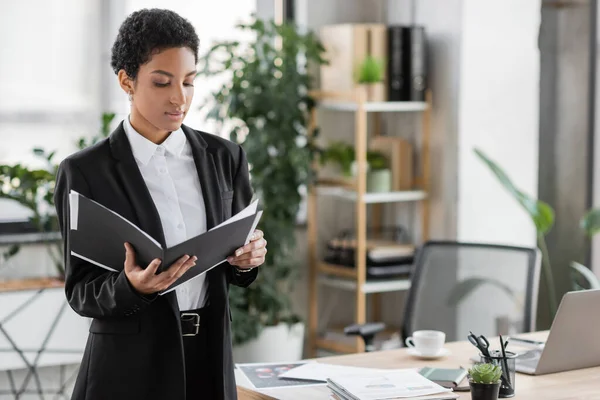 Femme d'affaires afro-américaine en costume noir regardant le dossier avec des papiers tout en restant au bureau — Photo de stock