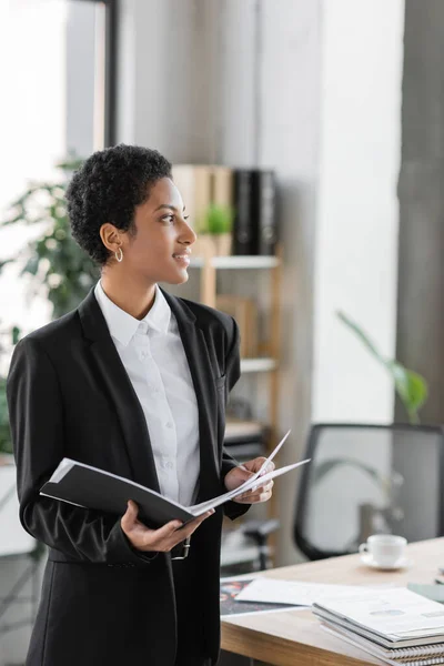 Joyful african american businesswoman holding folder with papers and looking away in office — Stock Photo