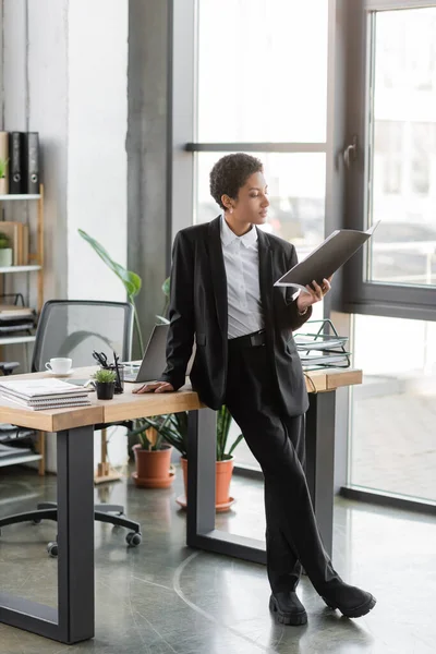 Повна довжина афроамериканської бізнес-леді в чорному костюмі, що стоїть з документами біля робочого столу в сучасному офісі — стокове фото