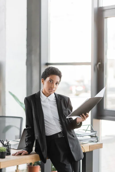 Jovem mulher de negócios afro-americana segurando pasta e olhando para a câmera perto da mesa de trabalho no escritório — Fotografia de Stock