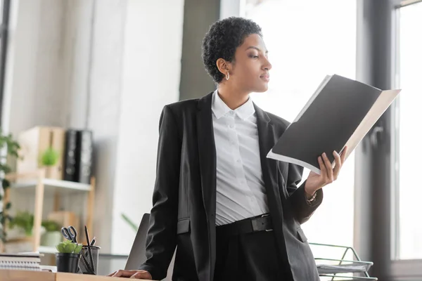 Elegante mujer de negocios afroamericana en blazer negro leyendo documentos en carpeta mientras está de pie en la oficina - foto de stock