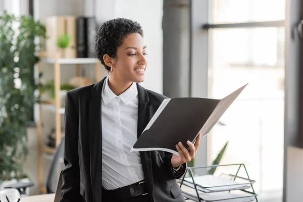 Pleased african american businesswoman in black blazer looking at documents in folder while standing in office — Stock Photo
