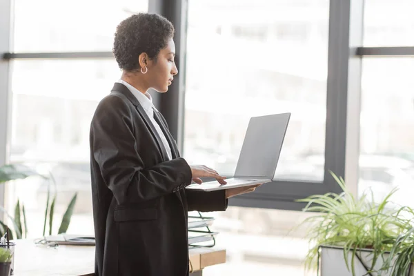 Vista lateral de la mujer de negocios afroamericana en traje negro usando computadora portátil con pantalla en blanco mientras está de pie en la oficina - foto de stock