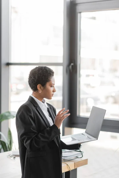 Visão lateral da empresária afro-americana em blazer preto acenando a mão durante chamada de vídeo no laptop no escritório — Fotografia de Stock