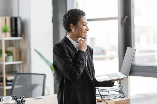 Side view of happy african american businesswoman holding laptop with blank screen while standing in office — Stock Photo