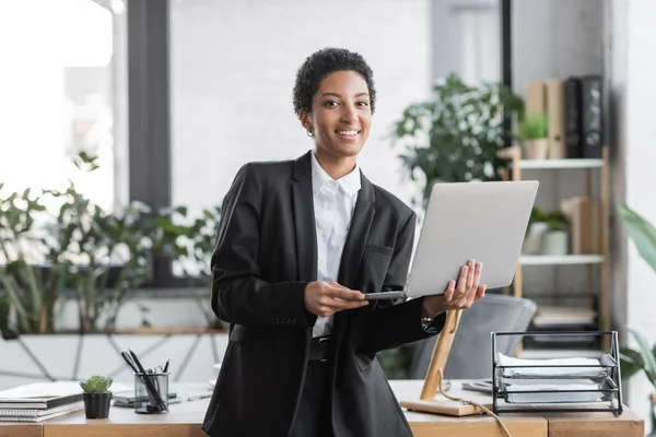 Alegre y elegante mujer de negocios afroamericana de pie con el ordenador portátil y sonriendo a la cámara en la oficina — Stock Photo