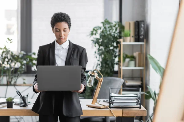 Африканский американский менеджер в черном блейзере стоит с ноутбуком рядом с рабочим столом в современном офисе — стоковое фото