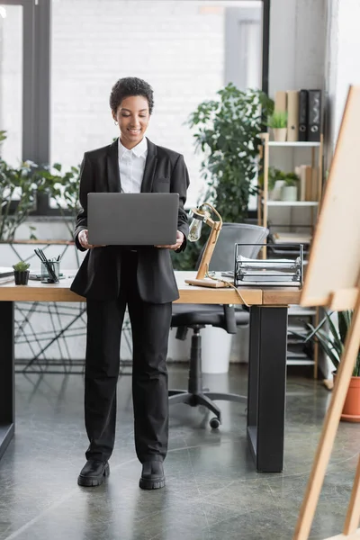 Повна довжина успішної афроамериканської бізнес-леді в чорному костюмі тримає ноутбук, стоячи на робочому столі в офісі — стокове фото