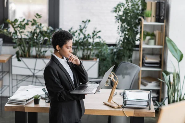 Vue latérale d'une femme d'affaires africaine américaine réfléchie debout dans un bureau moderne et regardant un ordinateur portable — Photo de stock