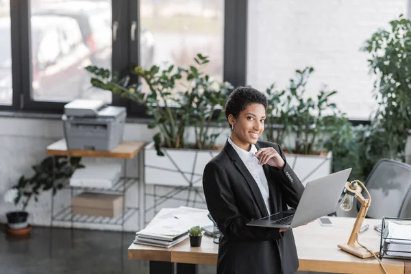 Alegre mujer de negocios afroamericana mirando a la cámara mientras está de pie con el ordenador portátil cerca del escritorio en la oficina moderna - foto de stock