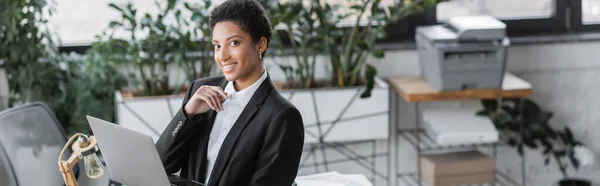 Complacida mujer de negocios afroamericana en chaqueta negra sosteniendo el ordenador portátil y sonriendo a la cámara en la oficina contemporánea, pancarta - foto de stock