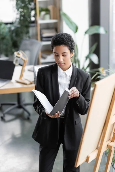 Femme d'affaires afro-américaine en costume noir regardant les documents dans le dossier tout en se tenant près du tableau de note dans le bureau — Photo de stock