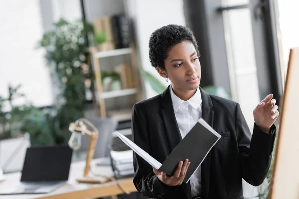 Elegante mujer de negocios afroamericana sosteniendo documentos y señalando borrosa pizarra en la oficina - foto de stock