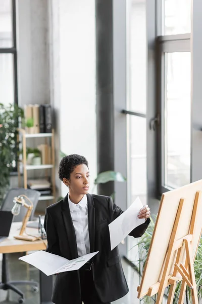Jovem empresária afro-americana segurando papéis e olhando para o quadro de notas enquanto trabalhava no escritório — Fotografia de Stock