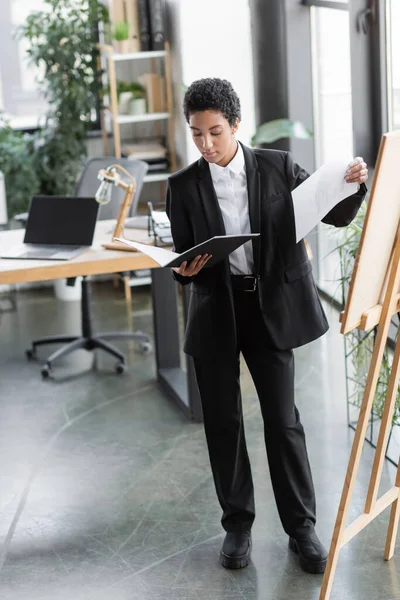 Comprimento total da empresária afro-americana de terno preto em pé com documentos perto do quadro de notas no escritório contemporâneo — Fotografia de Stock