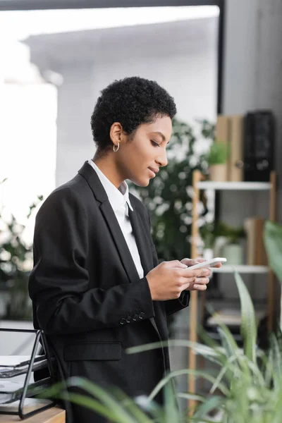 Vista lateral de la mujer de negocios afroamericana en la mensajería de desgaste formal negro en el teléfono móvil en la oficina - foto de stock