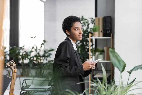 Vista laterale della donna d'affari africana americana sorridente in giacca nera che tiene lo smartphone e distoglie lo sguardo in ufficio — Foto stock