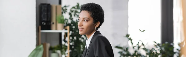 З іншого боку, молода афроамериканська бізнесменка в чорному блейзері посміхається і озирається в офіс, банер — стокове фото