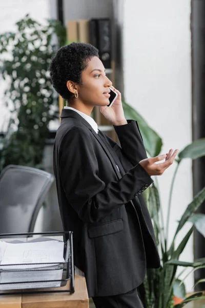 Vista lateral del gerente afroamericano en blazer negro gestos durante la conversación en el teléfono móvil en la oficina - foto de stock