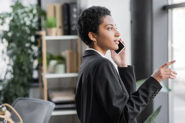 Vista lateral de la joven mujer de negocios afroamericana americana en chaqueta negra señalando con el dedo y hablando por teléfono celular en la oficina - foto de stock