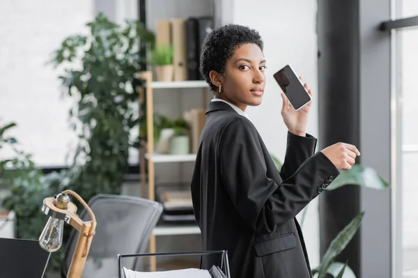 Elegante mujer de negocios afroamericana sosteniendo teléfono inteligente con pantalla en blanco y mirando a la cámara en la oficina moderna - foto de stock