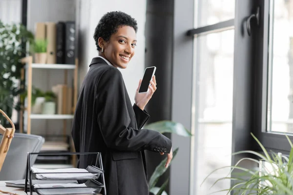 Freudige afrikanisch-amerikanische Geschäftsfrau im schwarzen Blazer blickt in die Kamera, während sie ihr Smartphone mit leerem Bildschirm im Büro hält — Stockfoto
