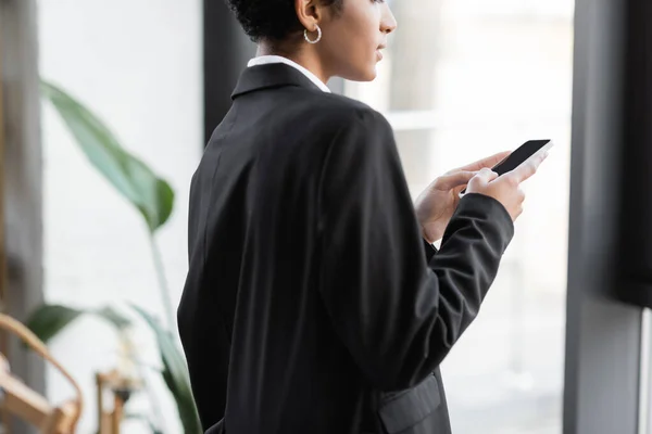 Частковий вигляд молодої афроамериканської бізнес-леді в чорному блейзер чаті на мобільному телефоні в офісі — стокове фото