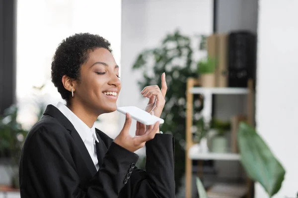 Cheerful african american businesswoman holding smartphone with white screen while sending voice message in office — Stock Photo